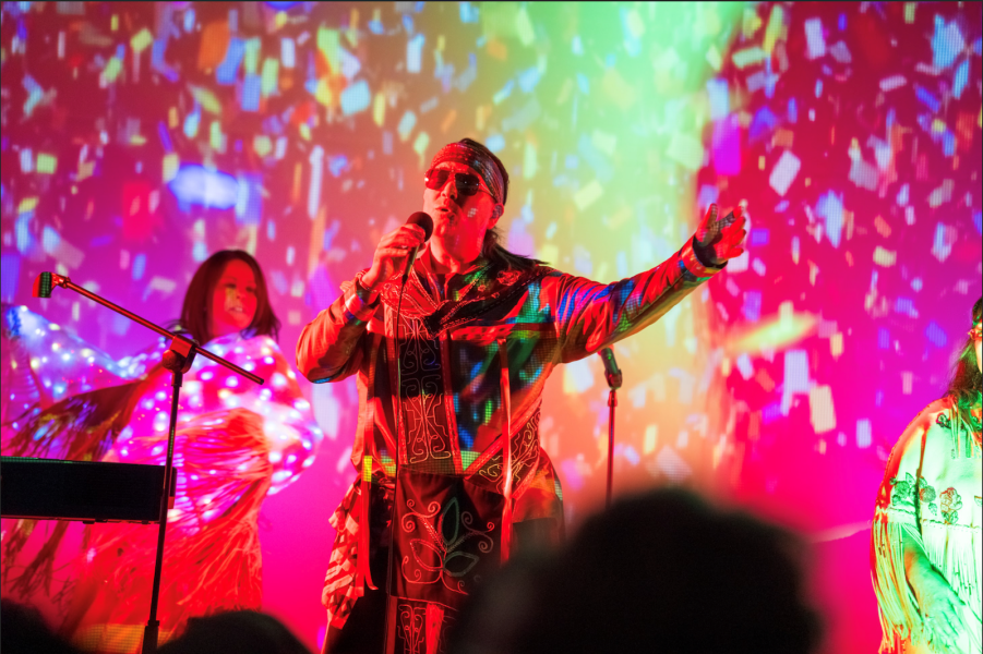 A singer in colorful, patterned clothing and sunglasses performs surrounded by vibrant multicolored lights and abstract patterns.