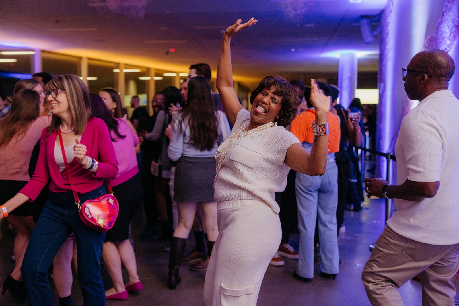 A middle-aged person in a white sweater dress and shoulder-length dark hair dances in a colorfully lit crowd
