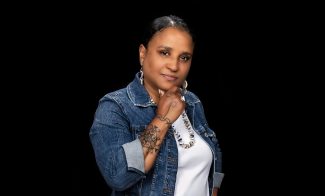 A woman stands against a black background, wearing a denim jacket over a white shirt and a red patterned skirt with white and orange bands. She has tattoos, jewelry, and her hair pulled back