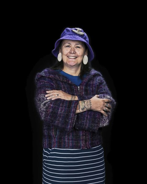 A smiling person stands with arms crossed, wearing a purple hat, patterned sweater, and striped skirt against a black background. They have small, circular facial tattoos and tattoos on their arms