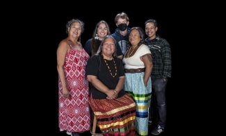 A group of six people, including four women in colorful dresses and two younger people in casual clothes, stands and sits together smiling against a plain black background