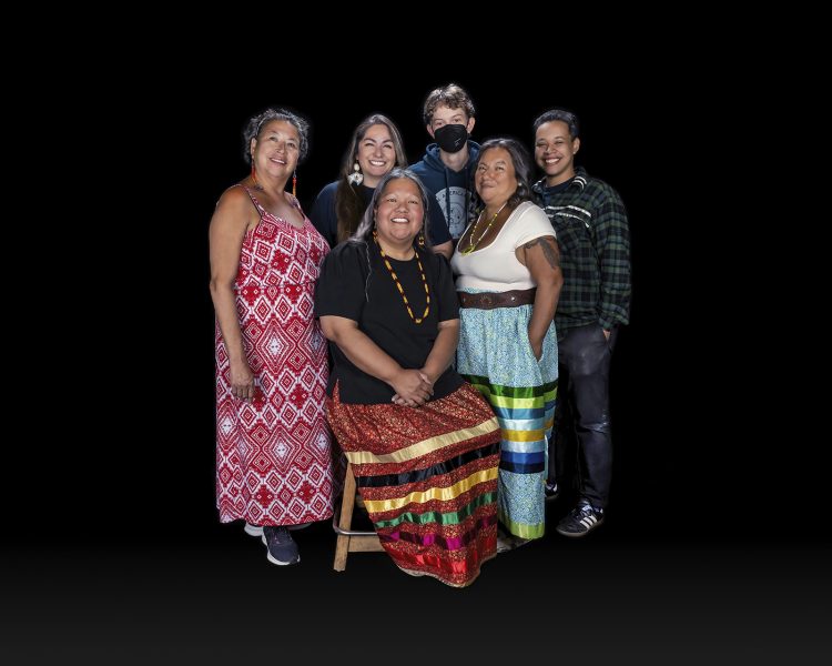 A group of six people, including four women in colorful dresses and two younger people in casual clothes, stands and sits together smiling against a plain black background