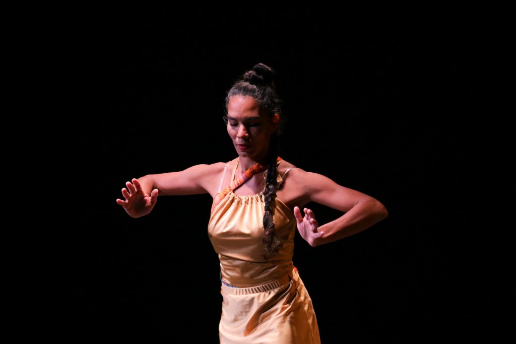 A woman in a yellow dress performs on a dark stage, her arms raised and bent at the elbows, with her long hair styled in a braid.