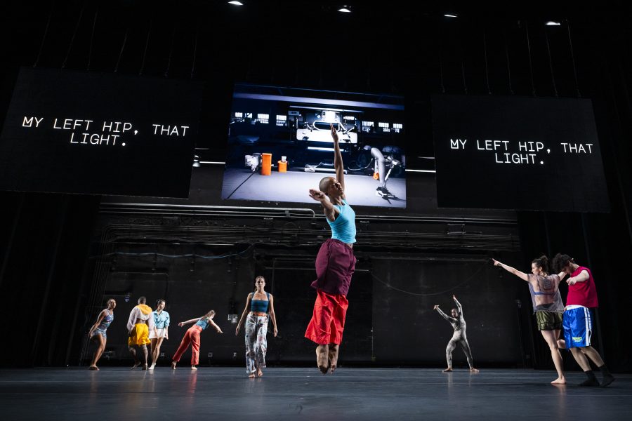 A group of dancers from Rashaun Mitchell + Silas Riener: Open Machine perform energetically on stage, with two large screens above displaying the text “MY LEFT HIP, THAT LIGHT” and an image of a person with equipment between them.