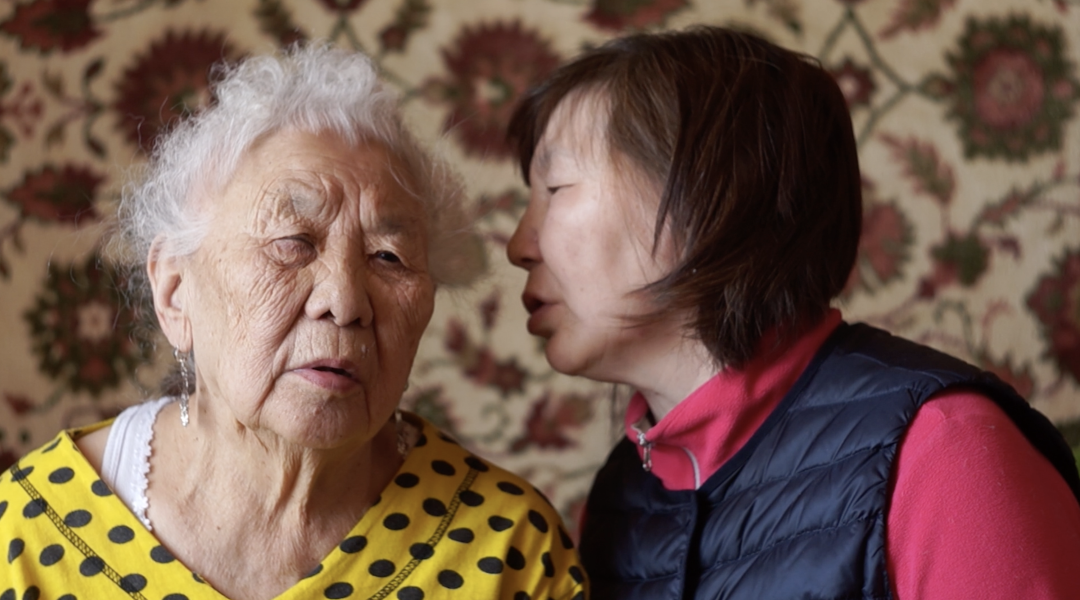 An elderly woman in a yellow polka-dot top sits next to a younger woman whispering in her ear, set against a floral-patterned tapestry