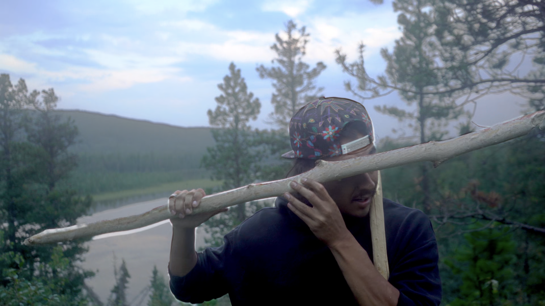 A person wearing a colorful baseball cap holds a large, pale tree branch over their shoulder, standing in a forested area.