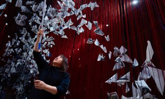 A woman reaches up with a pole toward many suspended paper birds and photographs, hanging among numerous red strings from the ceiling in an artistic installation.
