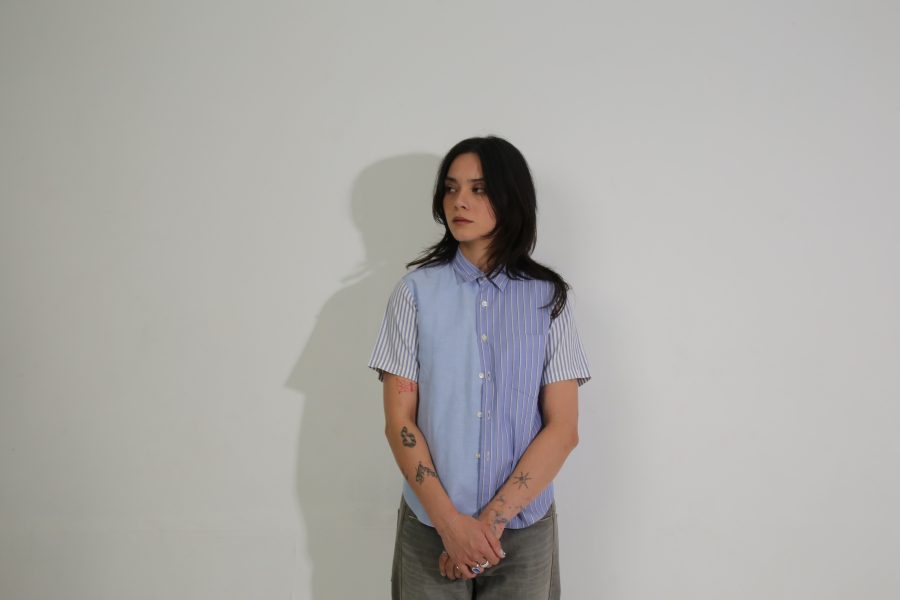 A person with long dark hair, wearing a short-sleeved blue and white striped shirt and gray pants, stands against a plain white wall.