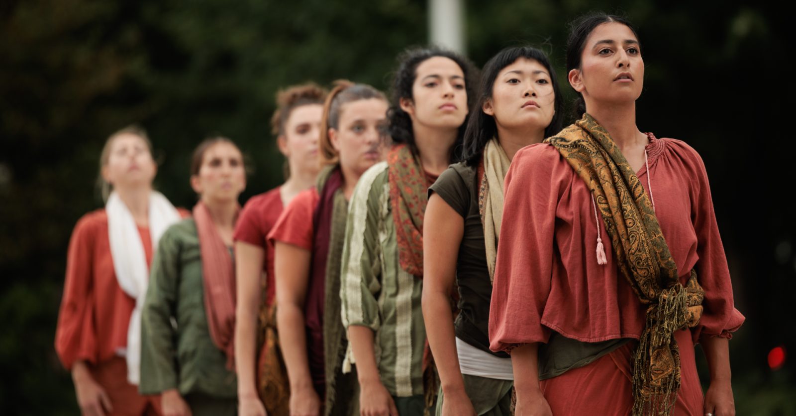 Seven performers, wearing earth-toned clothing and scarves, looking forward with serious expressions.
