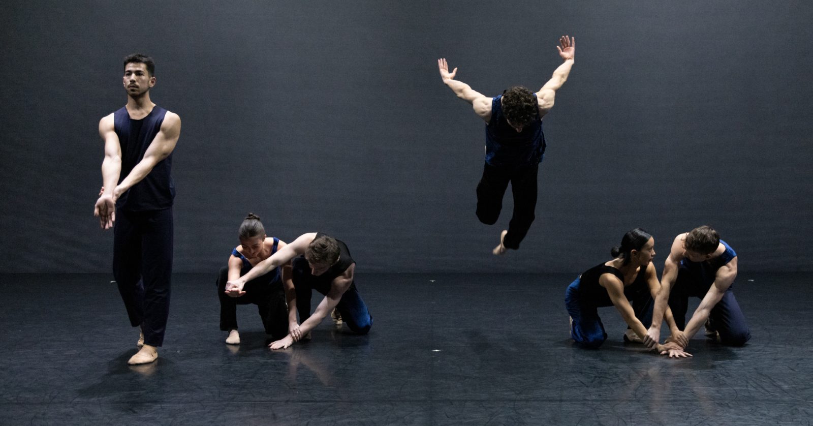 Six dancers perform on a dark stage; one stands alone left, two pairs kneel and interact right, while one dancer leaps dramatically in mid-air at center, arms raised, against a simple black background.
