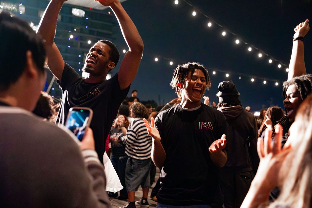 A group of people dance and celebrate outdoors at night under string lights. Two men in black ICA Teens shirts smile and raise their arms, surrounded by a lively crowd. One person holds up a phone to capture the moment.