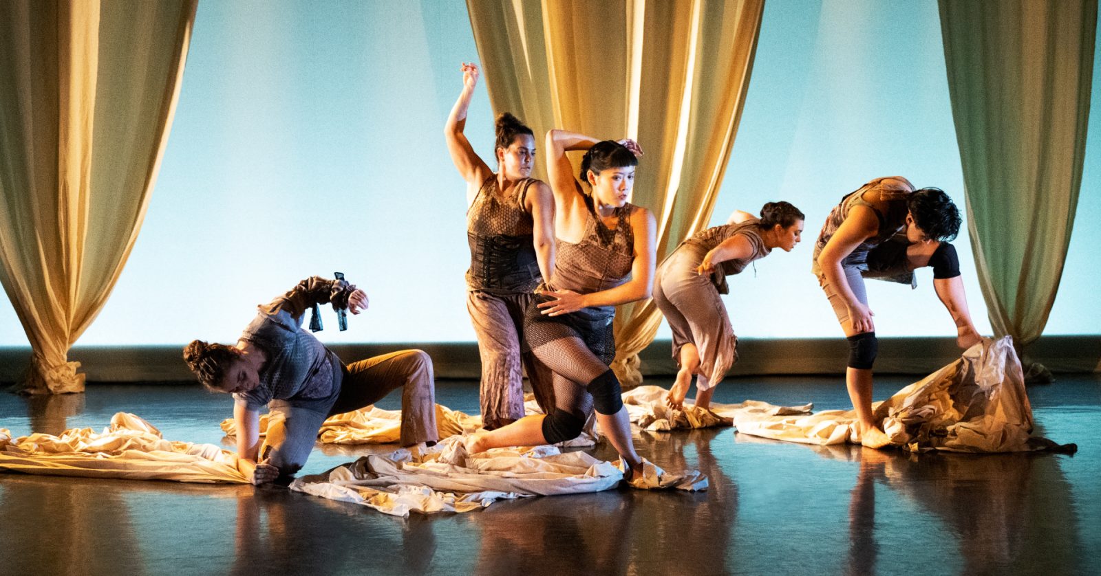 Five dancers in varied, expressive poses perform on stage among draped fabric and sheets, with warm lighting and sheer curtains in the background, creating a dynamic and dramatic scene.