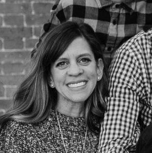 A woman with shoulder length hair and a sweater smiles against a brick wall