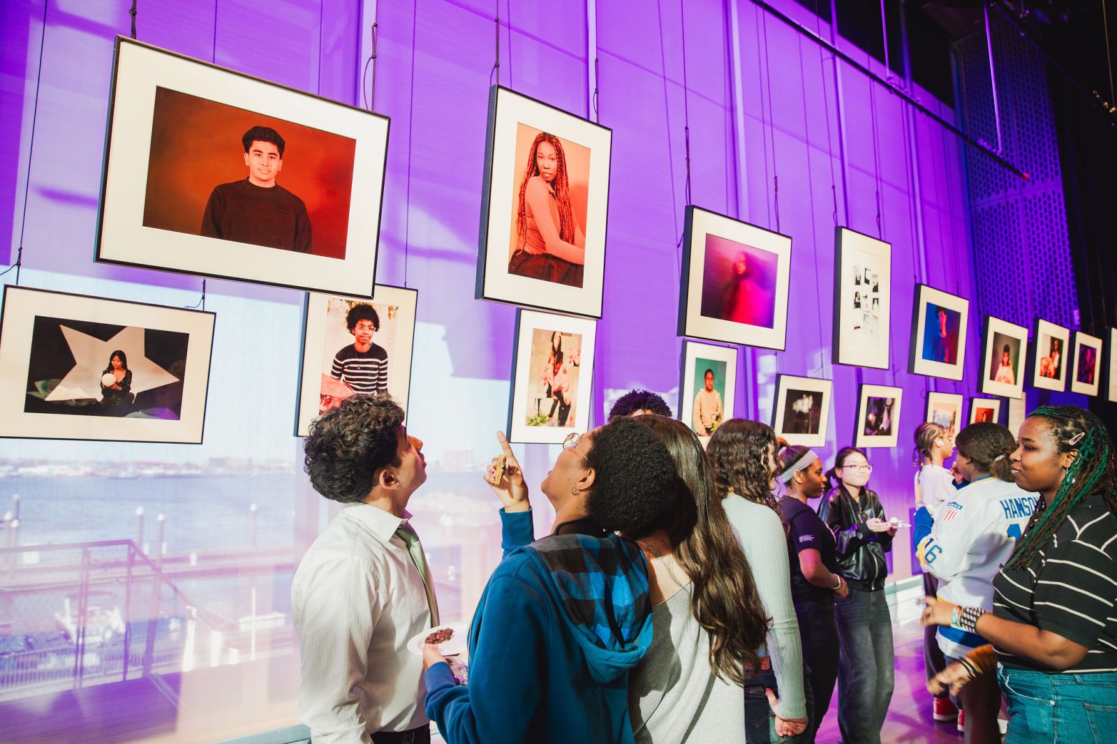 A group of people look at framed portraits hanging in a brightly lit gallery with purple-tinted walls and large windows showing water outside.