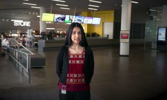 A woman with long dark hair wearing a red and black embroidered top stands indoors in a modern, spacious lobby area with a store and digital screens in the background.