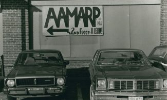 Two vintage cars are parked in front of a brick building. A sign on the wall reads AAMARP of NJ, 2nd floor-11 Leon with an arrow pointing left. The photo appears to be in black and white.