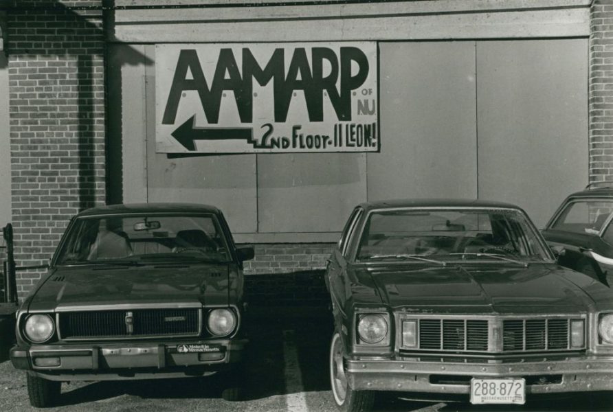 Two vintage cars are parked in front of a brick building. A sign on the wall reads AAMARP of NJ, 2nd floor-11 Leon with an arrow pointing left. The photo appears to be in black and white.