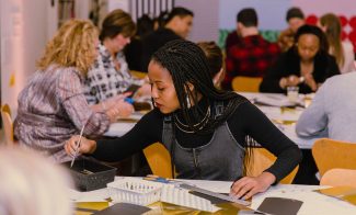 A young woman with braided hair sits at a table in a busy art classroom, focused on painting with a brush; other adults around her are also engaged in creative activities.