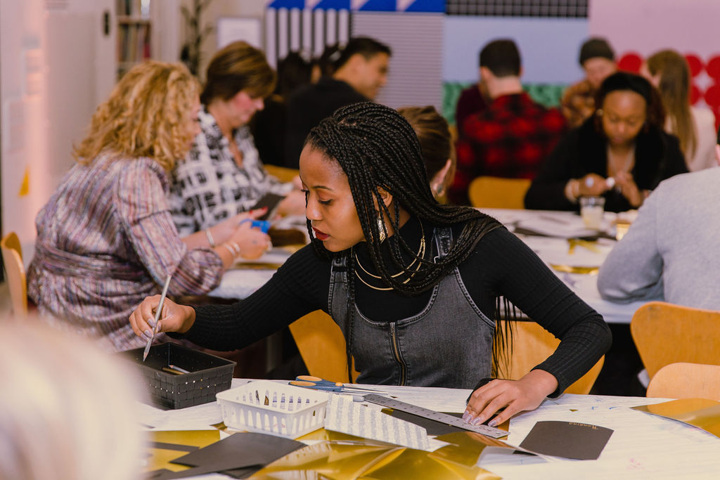 A young woman with braided hair sits at a table in a busy art classroom, focused on painting with a brush; other adults around her are also engaged in creative activities.