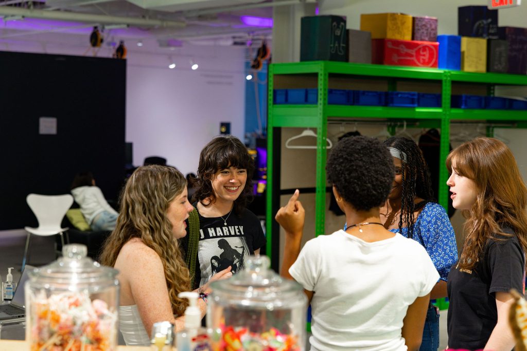 A group of five young people stand in a brightly lit room, talking and smiling. There are jars of candy in the foreground and colorful shelves in the background. The atmosphere appears friendly and lively.