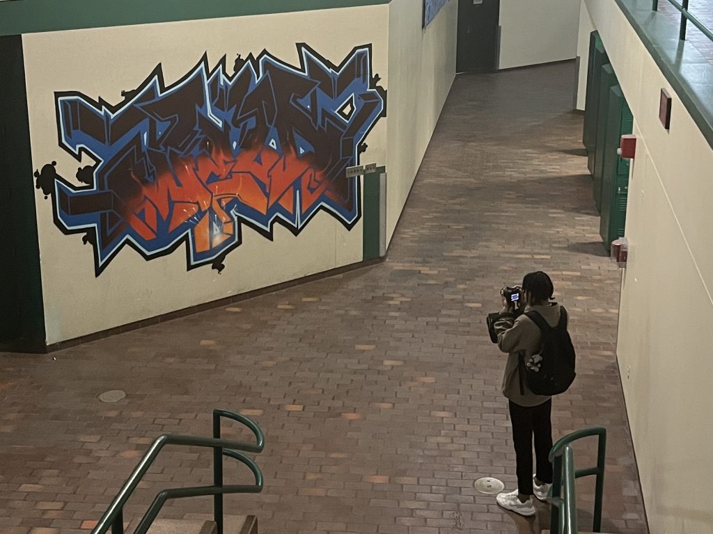 A person with a backpack photographs a colorful blue, black, and orange mural on an indoor wall in a building with brick flooring and green railings.