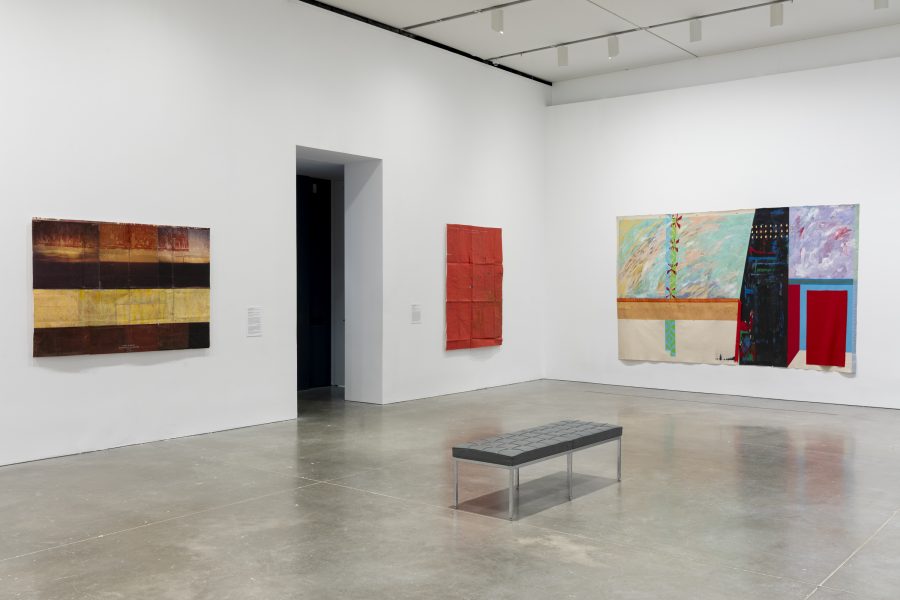 A gallery with three abstract paintings on white walls, a wide doorway, and a black bench on a polished concrete floor. The artworks feature bold colors, geometric shapes, and textured surfaces.