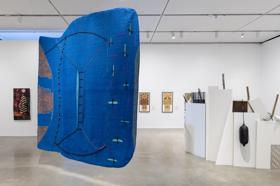 A gallery with a large, abstract blue textile sculpture suspended in the foreground, surrounded by artworks on white walls and a large sculptural installation on the floor