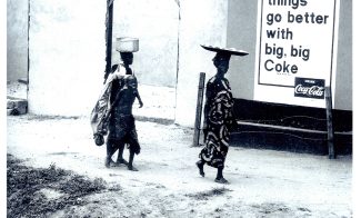 Two women and a child walk past a wall with a large Coca-Cola sign.