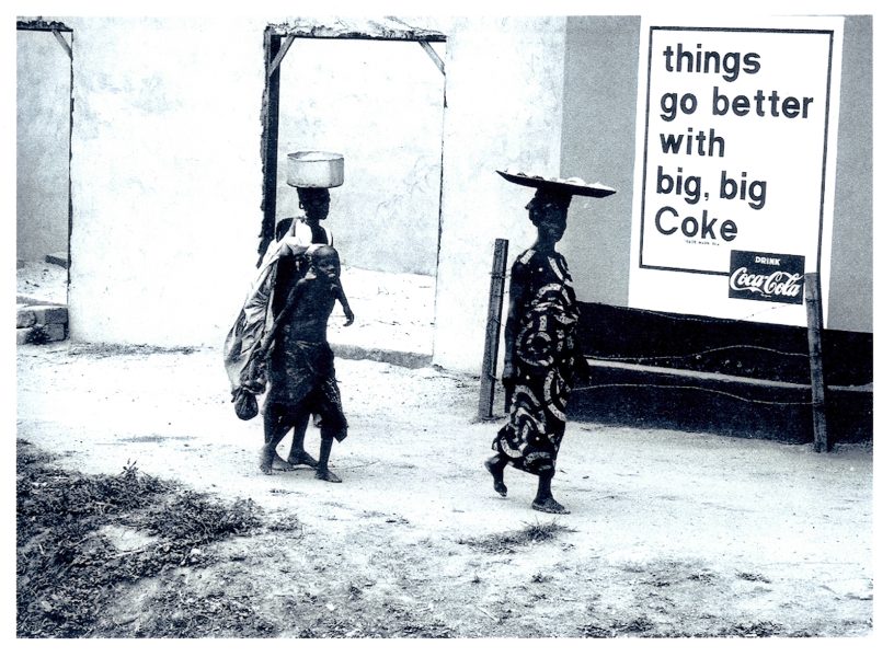 Two women and a child walk past a wall with a large Coca-Cola sign.