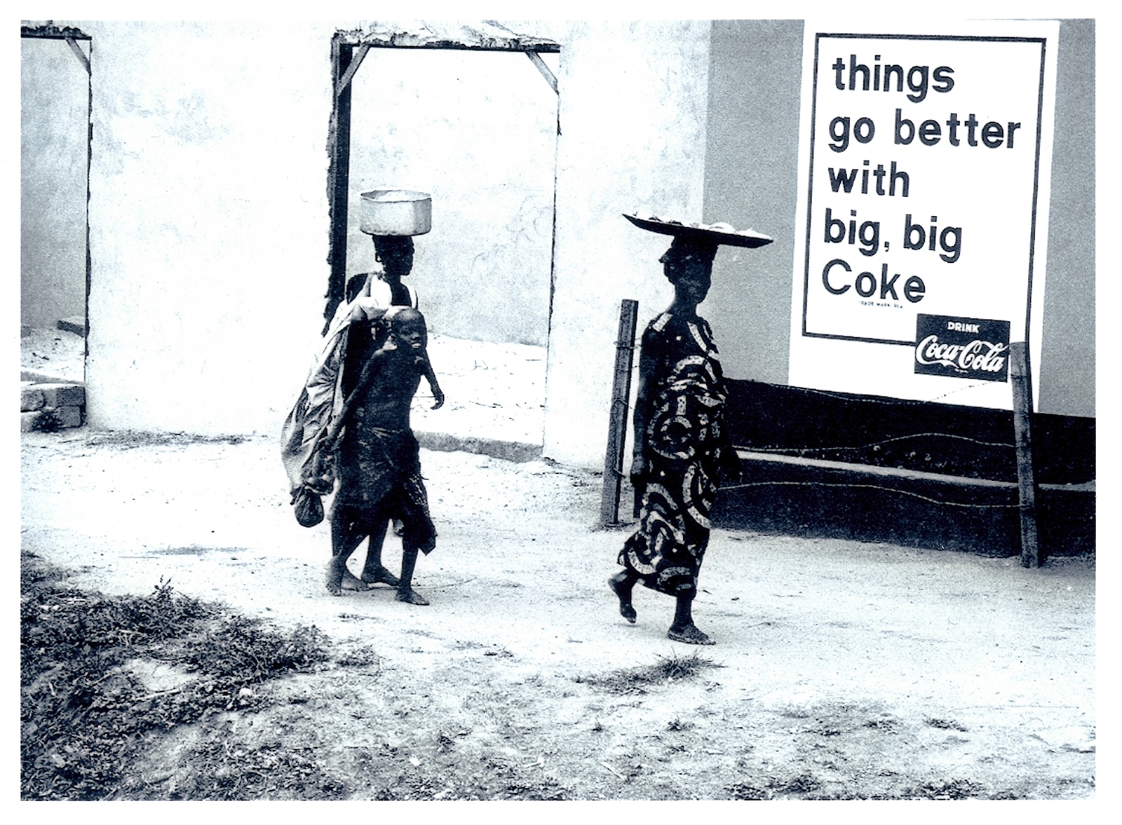 Two women and a child walk past a wall with a large Coca-Cola sign.