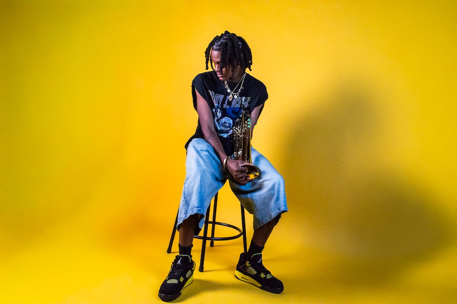 A person sits on a stool against a bright yellow background, looking down while holding a brass instrument.
