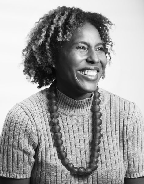 Black-and-white portrait of a woman smiling and looking slightly to the side. She has curly hair, wears a ribbed short-sleeve sweater, and a chunky beaded necklace. The background is plain and bright.