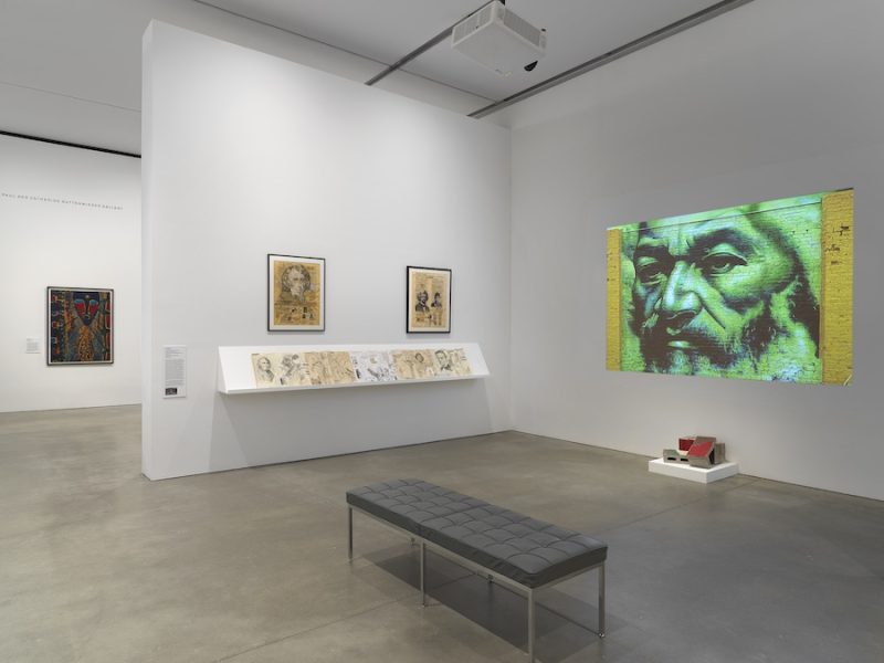 A modern art gallery at ICA The Artists' Voice features framed artwork on white walls, a display shelf with drawings, a gray bench, and a striking green-tinted projection of a bearded man above a small red-and-white sculpture.