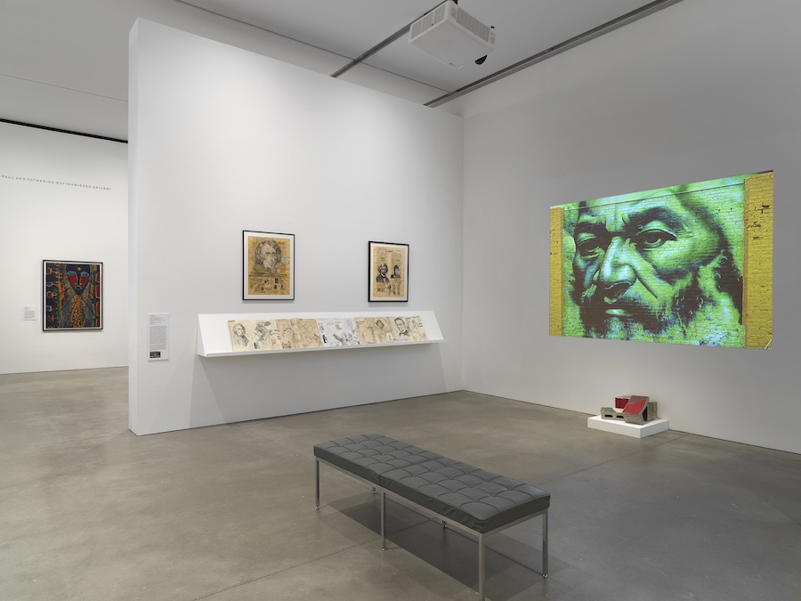 A modern art gallery at ICA The Artists' Voice features framed artwork on white walls, a display shelf with drawings, a gray bench, and a striking green-tinted projection of a bearded man above a small red-and-white sculpture.