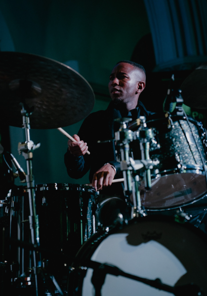 A drummer wearing dark clothing plays a drum set under moody stage lighting,