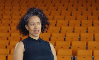 A woman with curly dark hair streaked with gray, wearing a sleeveless black top, sits in front of rows of empty orange auditorium seats. She is looking slightly to the side and smiling.