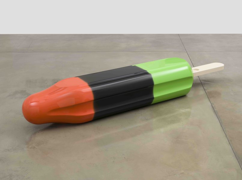A popsicle with three colored sections—orange, black, and green—lies on a concrete floor. The popsicle has a wooden stick and a glossy, reflective surface.