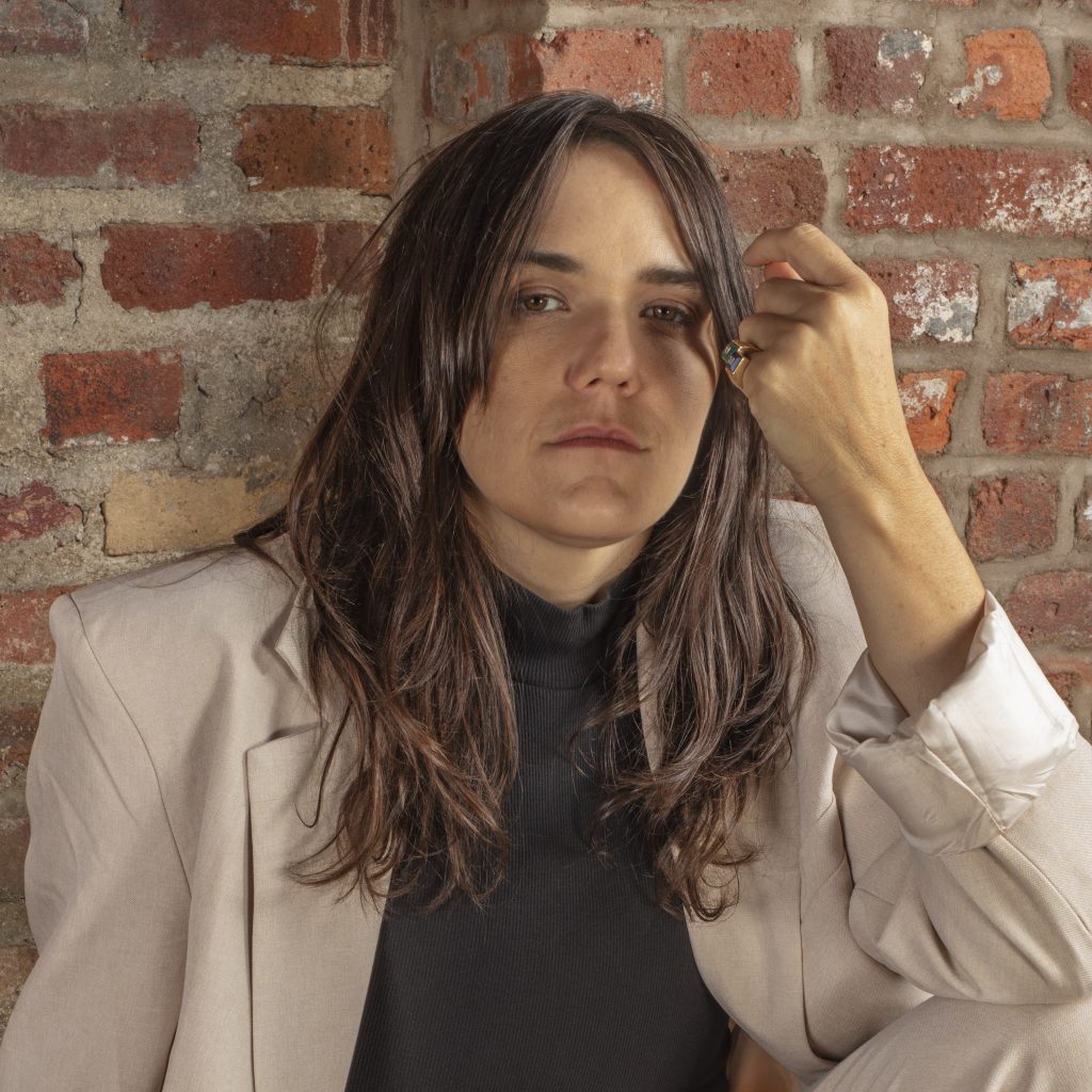 A person with long brown hair wearing a beige suit jacket and black top sits against a brick wall, looking at the camera with a serious expression and resting their hand near their face.