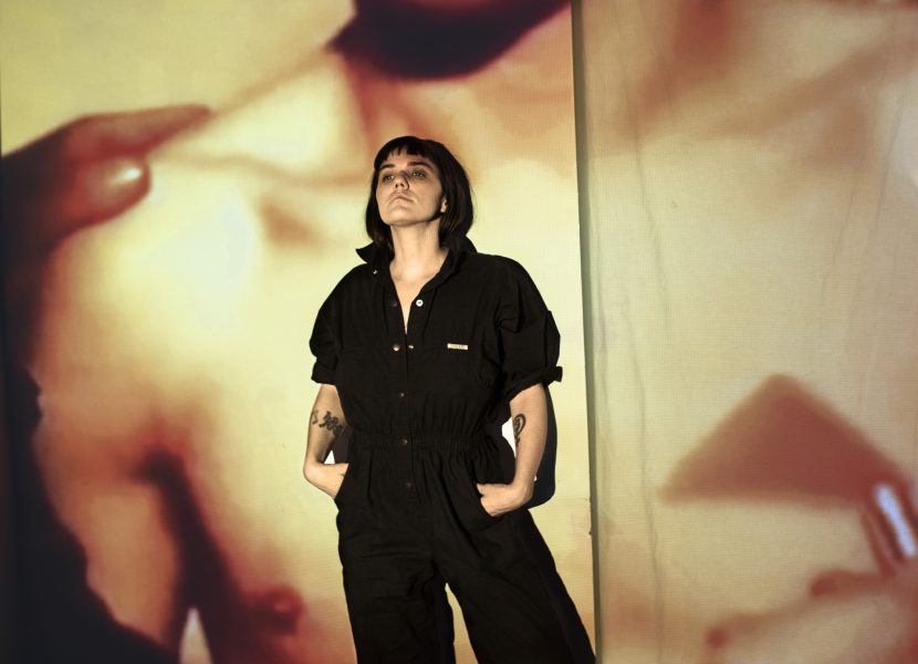 A person in a black jumpsuit stands confidently with hands in pockets, looking upward. Behind them, a large, blurred image of a bare-chested figure is projected onto a wall, creating a dramatic atmosphere.