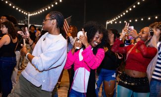 A group of people dance and celebrate under string lights at night. Two people in the center face opposite directions, smiling and dancing, while others take photos and enjoy the lively atmosphere around them.