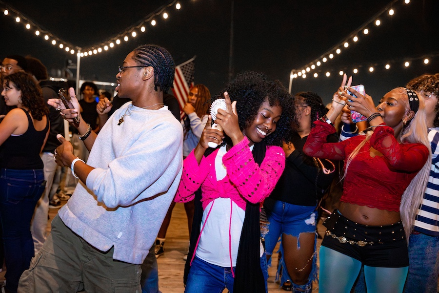 A group of people dance and celebrate under string lights at night. Two people in the center face opposite directions, smiling and dancing, while others take photos and enjoy the lively atmosphere around them.