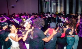 A large group of people form a conga line in a dimly lit room with purple lights, wearing matching black shirts with colorful lettering, appearing to celebrate and enjoy the festive atmosphere.