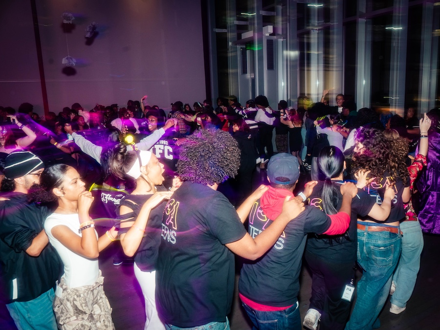 A large group of people form a conga line in a dimly lit room with purple lights, wearing matching black shirts with colorful lettering, appearing to celebrate and enjoy the festive atmosphere.