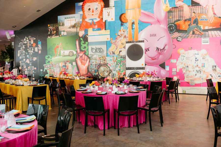 A colorful event space with round tables covered in bright pink and yellow tablecloths, black chairs, and vibrant table settings. In the background, a lively mural features playful, oversized cartoon and retro pop culture images.