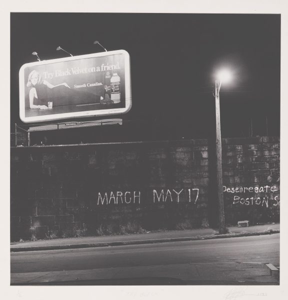 A black-and-white photo shows a street at night with a lit billboard advertising Black Velvet whisky above a dark stone wall. Graffiti on the wall reads MARCH MAY 17 and Desegregate BOSTON S. A streetlamp glows to the right.