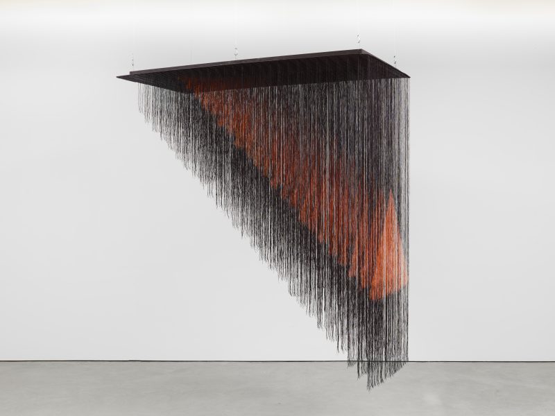 A suspended triangular installation made of hanging black strings forms a geometric shape, with a subtle orange-red gradient visible within the strands, set against a plain white wall and gray floor.