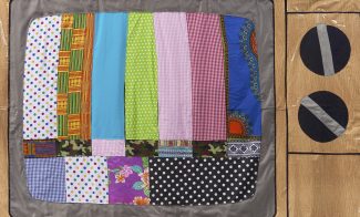 A handmade quilt designed to look like an old-fashioned TV displaying a colorful test pattern, reminiscent of an Opening Reception: Derrick Adams: View Master, with patchwork bars on the screen and two large black dials on the right side.