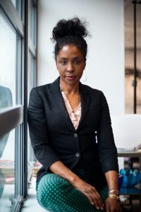 A woman with curly hair in a bun, wearing a black blazer, light pink blouse, and green polka-dot pants, sits by a window with her hands resting on her knee, looking confidently at the camera.