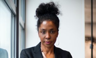A woman with curly hair in a bun, wearing a black blazer, light pink blouse, and green polka-dot pants, sits by a window with her hands resting on her knee, looking confidently at the camera.