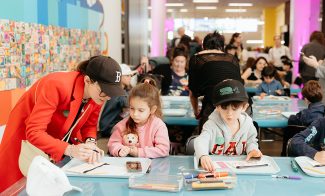 Adults and children sit at tables covered with art supplies, drawing and coloring. A woman in a red coat helps a young girl with brown hair, while a boy with a cap colors nearby. The room is busy and brightly lit.